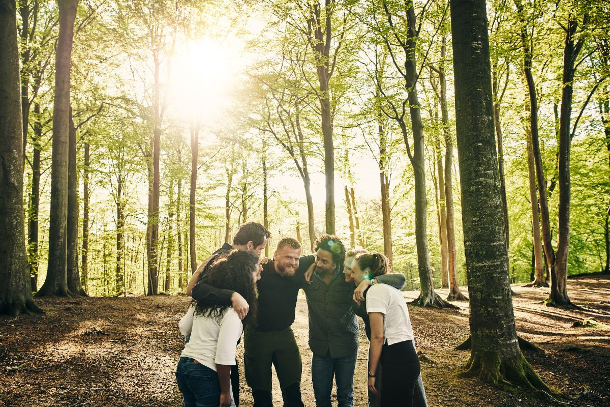 A group of colleagues stands in a circle, arms around each other, smiling and laughing in a forest setting during a corporate retreat. Sunlight filters through the trees, creating a warm and inviting atmosphere.